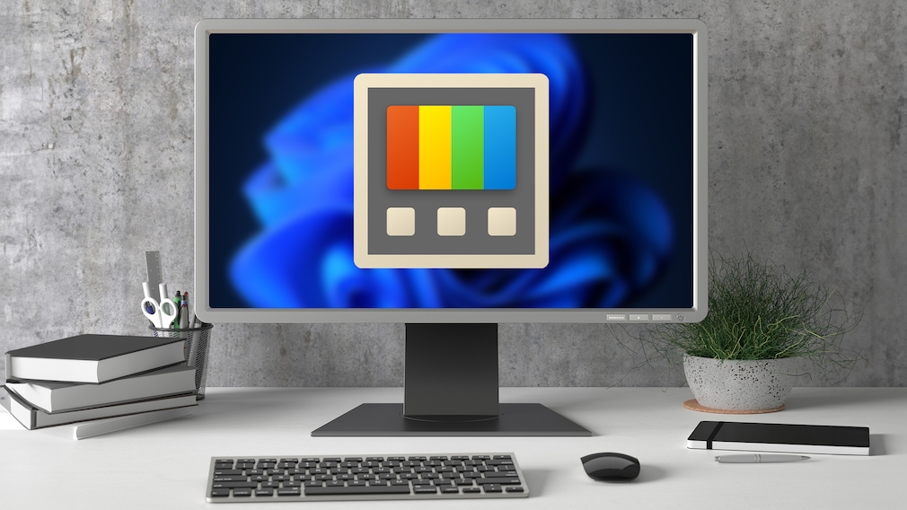 Ein Monitor steht auf einem Schreibtisch vor einer grauen Wand. Auf dem Bildschirm ist der blaue Windows-11-Hintergrund zu sehen, darüber befindet sich ein großes Symbol der Windows-Anwendung PowerToys mit vier farbigen Balken. Vor dem Monitor stehen eine Tastatur und eine Maus. Links liegen mehrere gestapelte Bücher sowie ein Stiftebecher mit Schere und Stiften. Rechts stehen eine Pflanze in einem Topf und ein flaches schwarzes Gerät mit einem Stift daneben.
