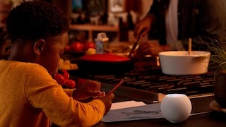 HomePod mini steht auf einem Tisch, daneben macht ein Kind Schulaufgaben.