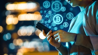 Mensch schaut aufs Handy, im Hintergrund Bitcoinsymbole