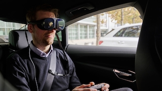 Ein Mensch sitzt mit einer VR-Brllle und einem Controller in einem Auto.