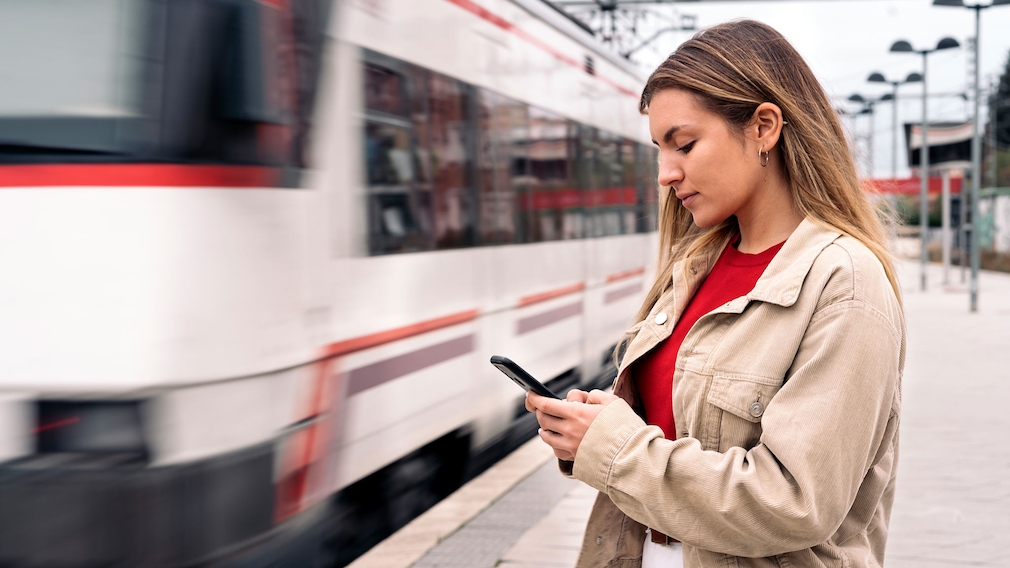 Eine Frau steht an einem Bahngleis und schaut auf ihr Handy.