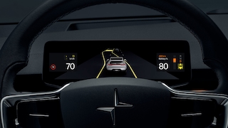 Hinterm dem Lenkrak zeigt ein Bildschirm die Rückansicht eines Autos und Farhzeugdaten an.
