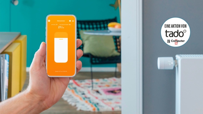 eine Hand hält ein Smartphone mit der Tado-App, im Hintergrund ein Wohnzimmer