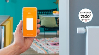 eine Hand hält ein Smartphone mit der Tado-App, im Hintergrund ein Wohnzimmer