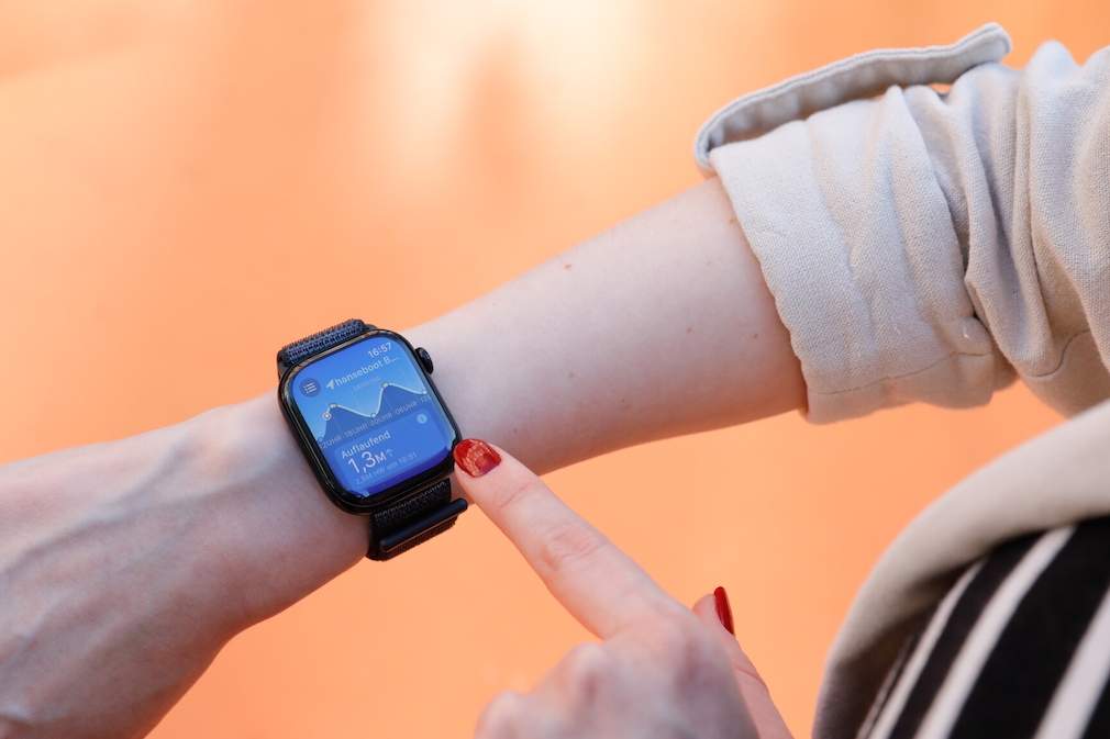 Apple Watch 10 am Handgelenk einer Person. Auf dem Display ist die Gezeiten-App mit aktuellen Werten zu sehen. Die Person deutet mit ihrem Zeigefinger auf das Display. Sie trägt roten Nagellack.