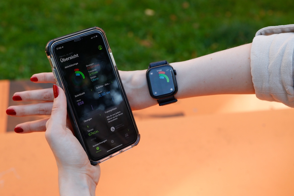 Apple Watch 10 am Handgelenk einer Person. Auf dem Display sind die Aktivitätsringe zu sehen. Daneben hält sie ein iPhone 11 mit der Fitness-App.