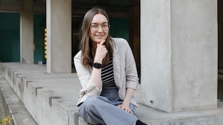 Eine Frau sitzt auf einem Betonuntergrund und hat das eine Bein auf dem anderen liegen. Sie stützt ihren rechten Arm auf dem Knie ab und trägt an dem Arm die schwarte Apple Watch 10. Sie lächelt in die Kamera.