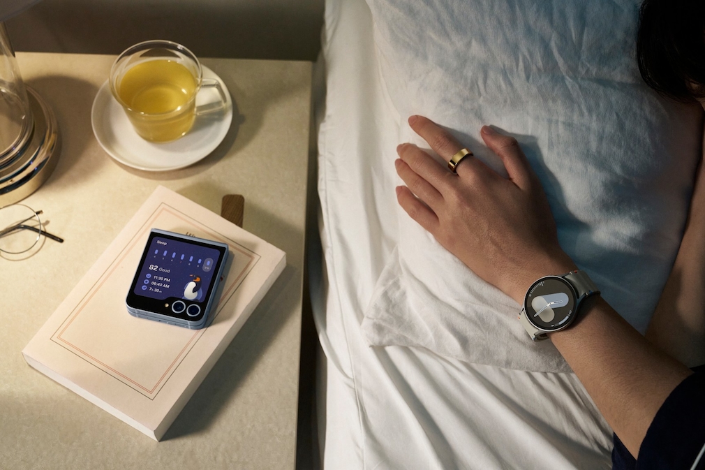 Arm liegt auf dem Bett mit einer Galaxy Watch 7 am Handgelenk und dem Galaxy Ring am Finger. Auf dem Tisch daneben liegt ein Galaxy Z Flip 6 auf einem Buch, daneben steht eine Tasse mit Tee