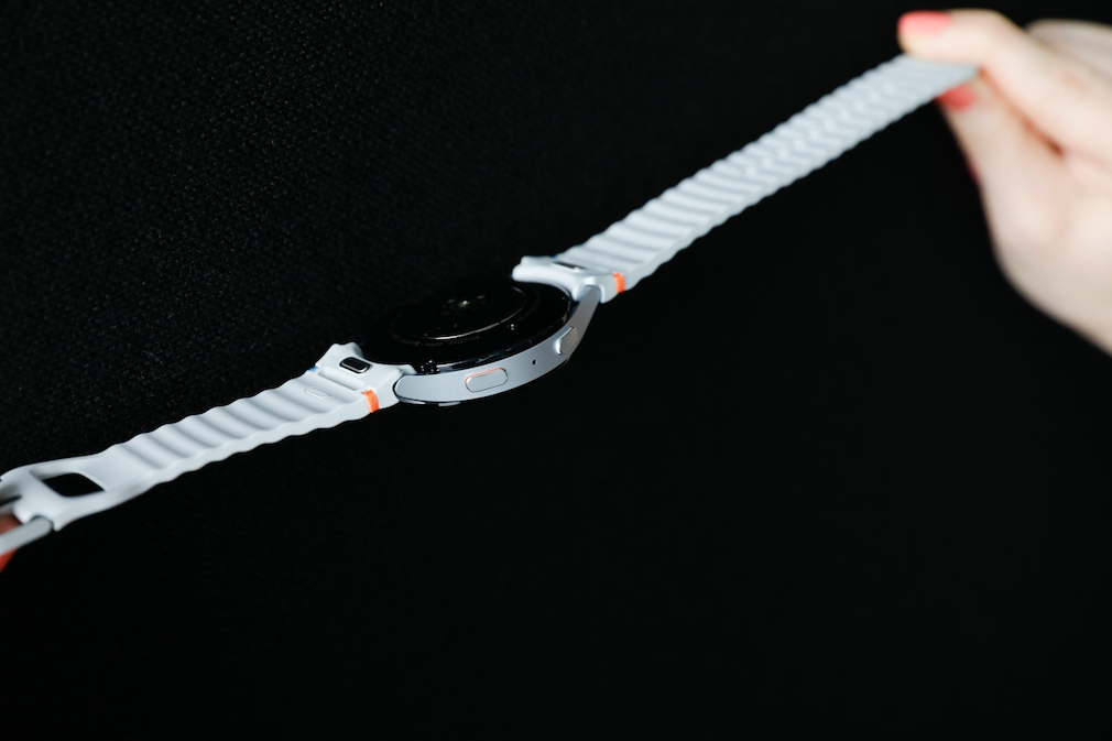 Galaxy Watch 7 gehalten von zwei Händen. Die Uhr ist mit der Rückseite nach oben gedreht und vor einem schwarzen Hintergrund platziert.