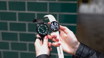 Zwei Hände halten die Samsung Galaxy Watch 6 Classic (links) und die Samsung Galaxy Watch 6 (rechts) vor einer Wand mit dunkelgrünen Backsteinen.