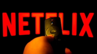 Netflix-Logo auf einem Fernsehbildschirm im Vordergrund ist eine Hand zu sehen, die eine Fernbedienung hält