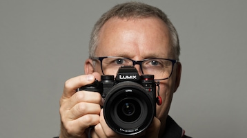 Testredakteur Sven Schulz hält die Panasonic Lumix S1IIE auslösebereit vor einem hellgrauen Hintergrund hoch. Die rechte Hand greift die Systemkamera am Griff, die linke Hand unterstützt von unten.