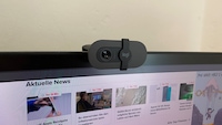 Die Webcam ist auf einem Monitor montiert. Links neben der Linse ist die „1080p“-Kennzeichnung erkennbar, die Abdeckung mit „logi“-Logo befindet sich mittig. Im Hintergrund zeigt der Bildschirm eine Nachrichtenwebseite.