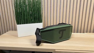 Der tragbare Bluetooth-Lautsprecher steht auf einem hellen Holztisch neben einem Pflanzgefäß. Das Gehäuse ist grün-schwarz, mit großem Teufel-Logo auf dem Frontgitter.