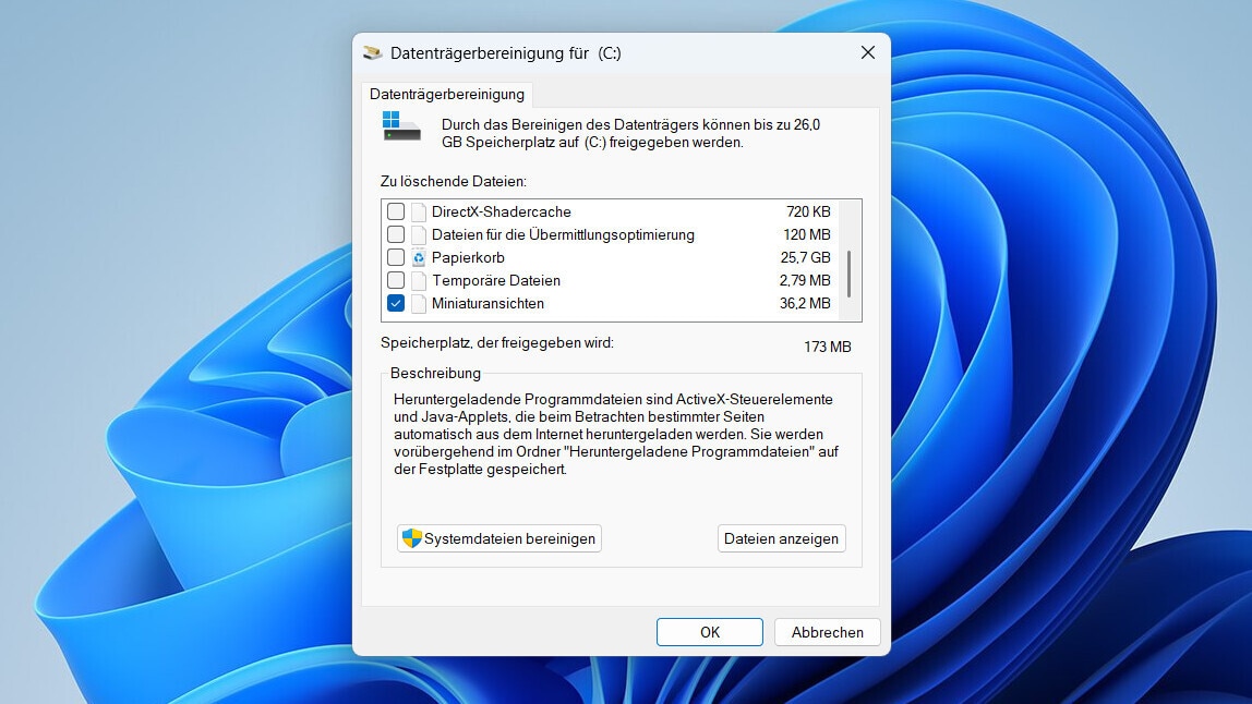 Screenshot eines Windows-11-Desktops mit geöffnetem Dialogfenster „Datenträgerbereinigung für (C:)“. Im Hintergrund ist der blaue Standardhintergrund von Windows 11 zu sehen. Im Fenster wird angezeigt, dass durch die Bereinigung des Laufwerks C: bis zu 26,0 GB Speicherplatz freigegeben werden können. Darunter befindet sich eine Liste mit auswählbaren Dateikategorien. Sichtbar sind unter anderem DirectX-Shadercache, Dateien für die Übermittlungsoptimierung, Papierkorb, Temporäre Dateien und Miniaturansichten. Neben den Einträgen stehen die jeweiligen Größen, darunter 120 MB, 25,7 GB, 2,79 MB und 36,2 MB. In der Liste ist Miniaturansichten ausgewählt. Unterhalb der Liste wird bei „Speicherplatz, der freigegeben wird“ der Wert 173 MB angezeigt. Im unteren Bereich befindet sich ein Beschreibungsfeld mit einem Text zu heruntergeladenen Programmdateien. Darunter sind die Schaltflächen „Systemdateien bereinigen“ und „Dateien anzeigen“ sichtbar. Rechts unten befinden sich die Schaltflächen „OK“ und „Abbrechen“.