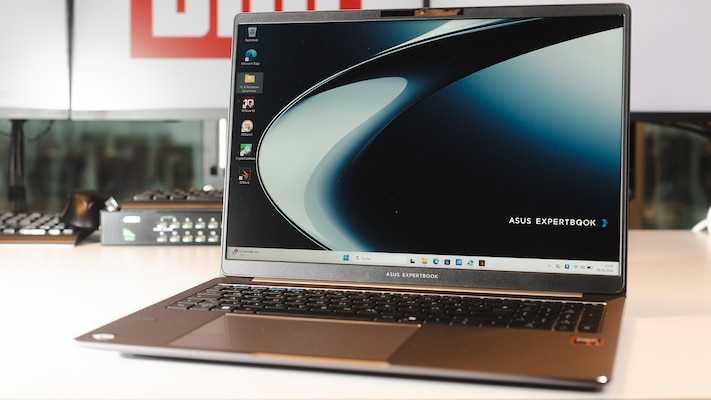 Aufgeklapptes Notebook auf einem Tisch, sichtbar sind Bildschirm mit Windows-Desktop, schwarze Tastatur, Touchpad und das graue Gehäuse, im Hintergrund stehen weitere Geräte auf einer Arbeitsfläche.