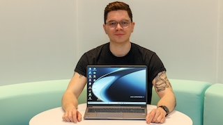 Frontale Aufnahme einer Person mit Brille, die hinter einem runden Tisch sitzt, vor ihr steht ein aufgeklapptes Notebook mit sichtbarem Bildschirm und Tastatur, im Hintergrund sind eine gepolsterte Sitzbank und eine helle Wand zu sehen.