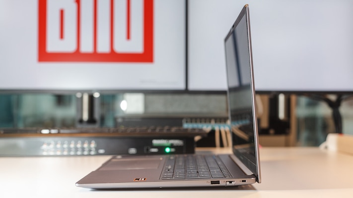 Seitliche Ansicht des aufgeklappten Asus ExpertBook PM3606CK auf einem Tisch, sichtbar sind das dünne Display, die Tastaturfläche und mehrere Anschlüsse an der rechten Seite, im Hintergrund stehen Monitore und weitere Geräte.