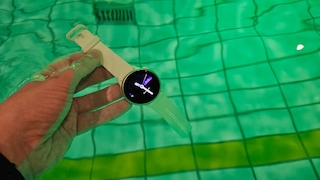 Eine Hand hält eine runde Smartwatch mit hellem Armband unter Wasser in einem Schwimmbecken. Das Display der Uhr ist eingeschaltet und zeigt ein analoges Zifferblatt. Das Wasser ist grünlich gefärbt, der Beckenboden mit Fliesen und Linien ist deutlich zu erkennen.