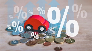 Auf einem Holztisch liegen viele Münzen verstreut. In der Mitte steht ein kleines rotes Spielzeugauto auf den Münzen. Darüber sind mehrere halbtransparente Prozentzeichen und große Pfeile nach oben eingeblendet, rechts dominiert ein sehr großes weißes Prozentzeichen.