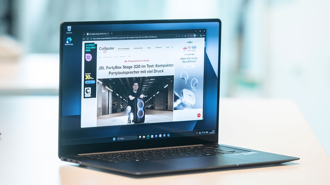 Ein aufgeklapptes Samsung Galaxy Book6 Pro steht auf einer hellen Tischfläche. Auf dem Display ist eine geöffnete Website zu sehen, die einen großen tragbaren Lautsprecher und eine Person daneben zeigt. Der Laptop ist schräg seitlich fotografiert, sodass Tastatur, Bildschirmrahmen und ein Teil des Gehäuses sichtbar sind. Der Hintergrund ist unscharf.