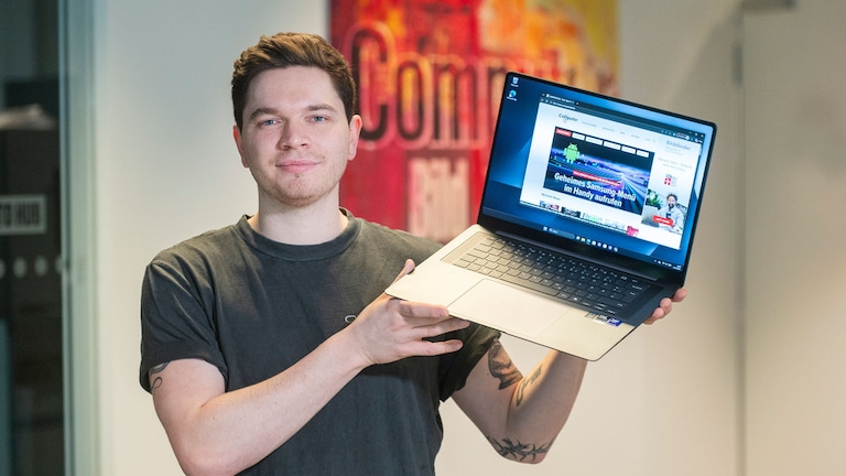 Eine Person hält ein geöffnetes Samsung Galaxy Book 6 Pro mit beiden Händen leicht schräg vor dem Körper. Auf dem Display ist eine geöffnete Website mit Technik Inhalten zu sehen. Der Hintergrund besteht aus einem unscharfen Innenraum mit einem farbigen Poster.