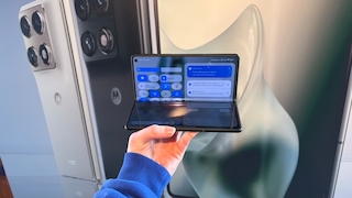 Eine Person hält ein aufgeklapptes faltbares Smartphone in der Hand. Auf dem Display ist ein Einstellungsmenü mit Schnellzugriffen und Benachrichtigungen zu sehen. Im Hintergrund stehen mehrere Smartphones mit sichtbaren Kameramodulen nebeneinander.