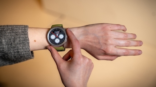 Nahaufnahme eines Handgelenks mit Smartwatch und grünem Armband. Eine zweite Hand berührt den Rand des runden Displays mit analogem Zifferblatt und drei kleinen Zusatzanzeigen. Hintergrund ist eine helle, einfarbige Fläche.