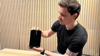 Eine Person hält einen schwarzen Sonos Era 100 Sl in den Händen und berührt die Oberseite. Im Hintergrund ist eine Wand mit vertikalen Holzleisten zu sehen.