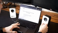Eine Person sitzt vor einem Laptop auf einem Schreibtisch und bedient mit der Hand einen kleinen weißen Logitech Z150 Lautsprecher. Auf dem Laptop ist eine Webseite geöffnet. Neben dem Laptop steht ein weiterer Lautsprecher.