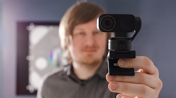 Nahaufnahme der Insta360 Link 2 Pro auf einem Halter, im Vordergrund scharf fokussiert. Das Objektiv mit Beschriftungsring ist deutlich sichtbar, daneben ein runder Sensor/LED-Bereich am Gehäuse. Im Hintergrund ist eine Person unscharf zu sehen, wodurch die Tiefenwirkung und der Fokus auf die Webcam betont werden.