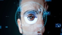 Alt-Text: Nahaufnahme eines menschlichen Gesichts vor dunklem Hintergrund. Im Mittelpunkt steht ein geöffnetes Auge, das direkt in die Kamera blickt. Über dem Auge sind mehrere halbtransparente, blau leuchtende grafische Elemente eingeblendet, darunter konzentrische Kreise, Markierungen, Fadenkreuze und rechteckige Informationsfelder. Die Grafiken sind kreisförmig um das Auge angeordnet und erinnern an eine digitale Benutzeroberfläche. Weitere kleine technische Anzeigen und Zahlen sind im Hintergrund verteilt sichtbar. Die Gesichtshälfte ist nur teilweise zu sehen, während das Auge den zentralen Bildbereich einnimmt.
