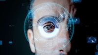 Alt-Text: Nahaufnahme eines menschlichen Gesichts vor dunklem Hintergrund. Im Mittelpunkt steht ein geöffnetes Auge, das direkt in die Kamera blickt. Über dem Auge sind mehrere halbtransparente, blau leuchtende grafische Elemente eingeblendet, darunter konzentrische Kreise, Markierungen, Fadenkreuze und rechteckige Informationsfelder. Die Grafiken sind kreisförmig um das Auge angeordnet und erinnern an eine digitale Benutzeroberfläche. Weitere kleine technische Anzeigen und Zahlen sind im Hintergrund verteilt sichtbar. Die Gesichtshälfte ist nur teilweise zu sehen, während das Auge den zentralen Bildbereich einnimmt.