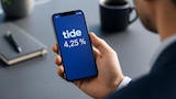 Eine Hand hält ein Smartphone, auf dessen Display der Schriftzug „tide“ und „4,25 %“ zu sehen sind; im Hintergrund liegen ein Notizbuch und eine Tasse.