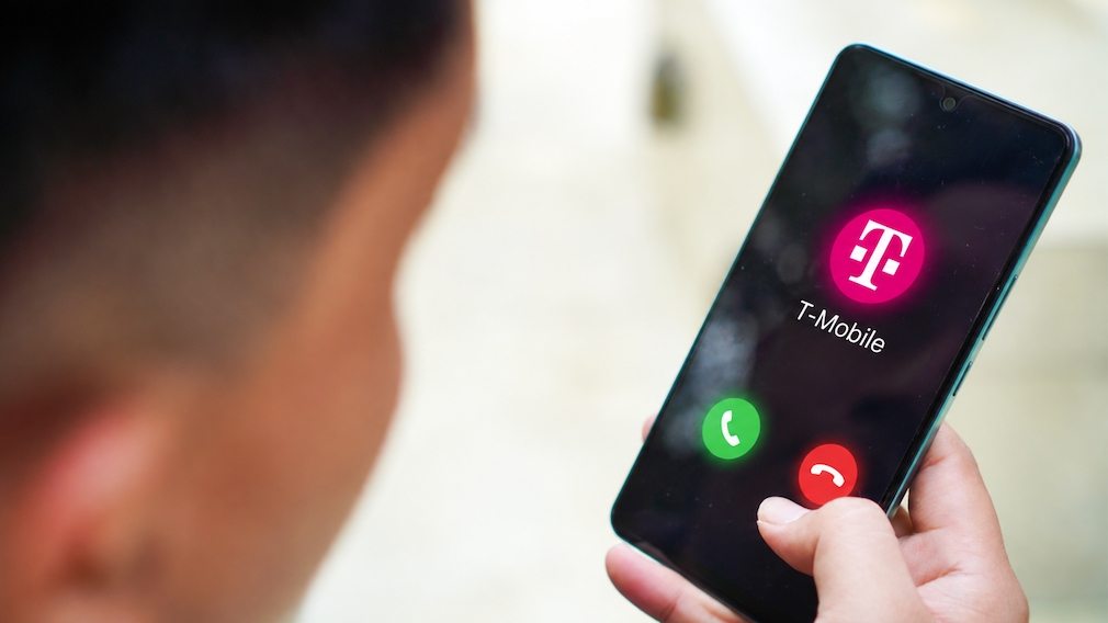 Person hält ein Smartphone in der Hand. Auf dem Display ist ein eingehender Anruf zu sehen. In der Mitte des Bildschirms befinden sich das magentafarbene T-Mobile-Logo und der Schriftzug „T-Mobile“. Darunter sind ein grünes Symbol zum Annehmen und ein rotes Symbol zum Ablehnen des Anrufs sichtbar. Der Kopf der Person ist unscharf im linken Vordergrund zu sehen, der Hintergrund ist ebenfalls unscharf.