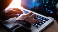 Nahaufnahme von zwei Händen auf der Tastatur eines aufgeklappten Laptops. Die Tasten werden von bläulichem Licht beleuchtet. Auf dem Bildschirm sind unscharfe blaue Linien und Zeichen auf dunklem Hintergrund zu sehen. Die Aufnahme ist in dunkler Umgebung entstanden, mit einem warmen Lichtreflex im linken oberen Bildbereich.
