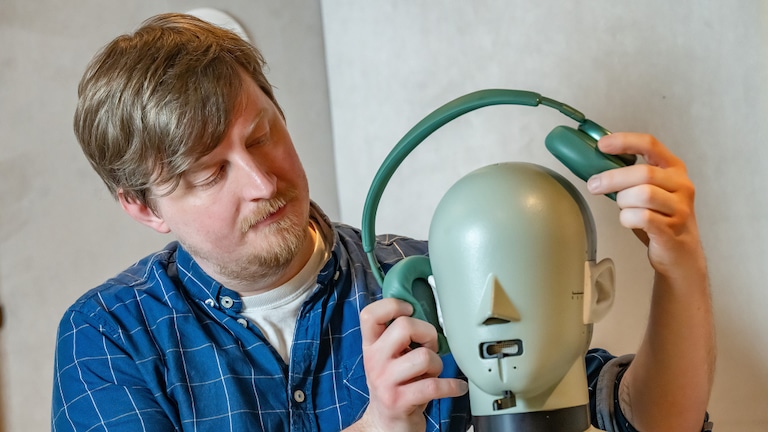 Person hält einen grünen Over-Ear-Kopfhörer an einen Kopf-/Kunstkopf für Messungen. Die Person steht links im Bild, der Kunstkopf ist rechts im Vordergrund. Hintergrund ist neutral, ohne erkennbare Markenbeschriftungen im Fokus.