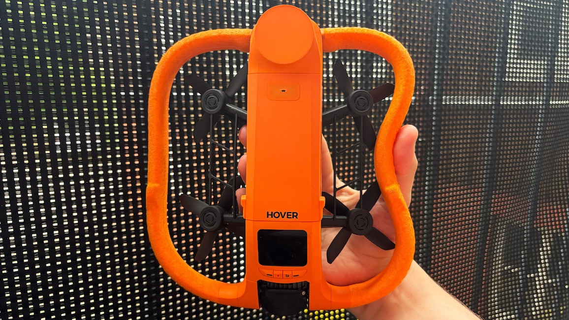 Eine Hand hält die orangefarbene Hoverair Aqua mit gebogenem Schutzrahmen vor einem dunklen Gitter.