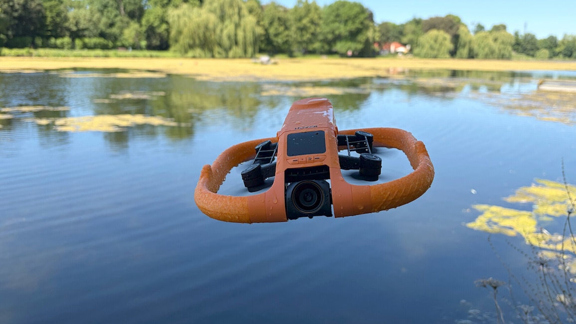 Eine orangefarbene Hoverair Aqua fliegt über einem See. Vorn ist die Kamera zu sehen, im Hintergrund spiegeln sich Bäume im Wasser.