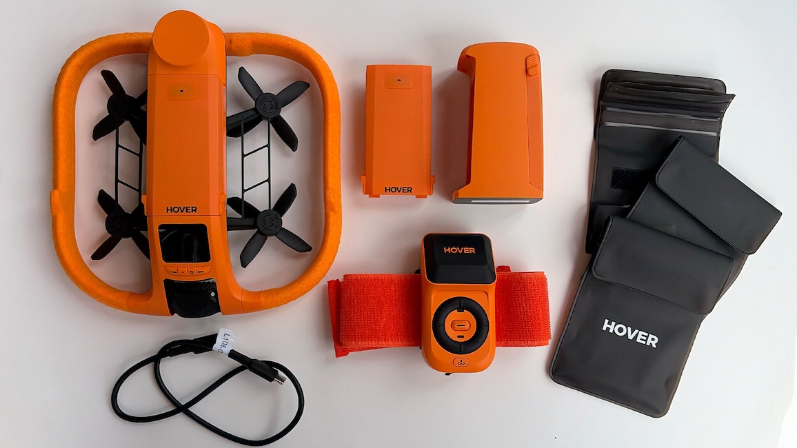 Links liegt die orangefarbene Hoverair Aqua mit schwarzem Propellerrahmen. Darunter liegt ein schwarzes Ladekabel. Oben mittig liegen zwei orangefarbene Akkus oder Bauteile. Unten mittig liegt die Lighthouse-Fernsteuerung mit rotem Band. Rechts liegen schwarze wasserdichte Akku-Taschen.