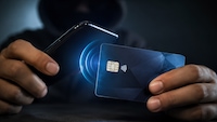 Nahaufnahme von zwei Händen, die ein Smartphone und eine kontaktlose Zahlungskarte halten. Das Smartphone befindet sich links im Bild und ist leicht zur Karte geneigt. Die Karte ist dunkelblau, zeigt einen sichtbaren Chip sowie ein Kontaktlos-Symbol und wird rechts gehalten. Zwischen Smartphone und Karte sind mehrere blaue, konzentrische Lichtkreise dargestellt. Im Hintergrund ist eine dunkel gekleidete Person unscharf zu sehen. Die Szene ist insgesamt dunkel ausgeleuchtet.