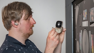 Person steht neben einem Mess-/Testaufbau (Bildschirm mit Testcharts). In einem Ohr steckt ein In-Ear, in der Hand ein offenes Case mit Display im Deckel. Die Person hält zusätzlich einen einzelnen In-Ear vor das Case, als Demonstration der Bedienung/Funktionen.