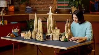 Eine Person sitzt an einem Tisch und betrachtet ein großes Modell der Sagrada Família aus Klemmbausteinen. Das Modell besteht aus mehreren hohen Türmen und Gebäudeteilen, die auf dem Tisch verteilt aufgebaut sind. Im Hintergrund sind ein Wohnraum mit Holzboden, Backsteinwand, Pflanzen und verschiedenen Möbelstücken zu sehen. Auf dem Tisch stehen außerdem ein Becher mit Werkzeugen oder Stiften, eine Kerze und weitere kleine Gegenstände.