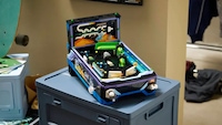 LEGO-Flipperautomat aus Klemmbausteinen auf einer blauen Kunststoffbox. Das Modell zeigt ein schwarzes Spielfeld mit grünen, weißen und blauen Elementen sowie zwei weißen Flippern im unteren Bereich. Im Hintergrund sind ein Teil eines Skateboards, Möbel und Kleidung in einem Raum zu sehen.