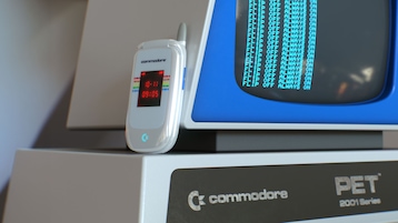 Weißes Commodore-Klapphandy mit kleinem Außendisplay lehnt an einem Monitor im Retro-Design. Auf dem Display des Handys sind Uhrzeit und weitere rote Anzeigen zu sehen. Daneben zeigt der Monitor mehrere Zeilen mit blauem Text auf schwarzem Hintergrund. Im Vordergrund ist ein Gehäuse mit Commodore-Logo und der Aufschrift "PET 2001 Series" sichtbar.
