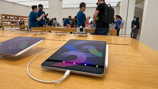 Ein iPad Air mit angeschlossenem Ladekabel und Eingabestift liegt auf einem Holztisch in einem Apple Store. Im Hintergrund beraten Mitarbeitende in blauen Shirts Kundinnen und Kunden an weiteren Produkttischen.
