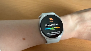 Samsung Galaxy Watch mit silbernem Gehaeuse und weissem Armband am linken Handgelenk. Auf dem Display ist unter dem Samsung-Health-Labs-Herzsymbol die Ueberschrift Etwas hoeher zu sehen, darunter eine farbige Skala mit einem Marker im oberen Bereich, der Hinweis Grundlinie und der Textanfang Deine Gefaessbelastung muss verbessert werden. Der Arm liegt schraeg auf einer hellen Holzflaeche.