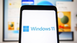Smartphone mit Windows-11-Logo vor Hintergrund mit Laptop mit Windows 11