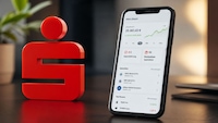 Auf einem Tisch steht links ein rotes Sparkassen-Symbol. Rechts daneben lehnt ein Smartphone, auf dessen Display eine Depotübersicht mit Depotwert, grüner Kursgrafik, „0 € Depotführung“, „0 € Kostenlose Sparpläne“ sowie ETF- und Aktienlisten zu sehen sind. Im unscharfen Hintergrund sind eine Pflanze, eine Lampe und ein Laptop erkennbar.