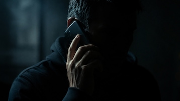 Das Bild zeigt eine Person, die in einer dunklen Umgebung mit einem Smartphone telefoniert. Das Gesicht ist größtenteils im Schatten verborgen, wodurch die Aufnahme anonym und geheimnisvoll wirkt. Die Beleuchtung ist sehr kontrastreich und konzentriert sich vor allem auf die Hand und das Smartphone.